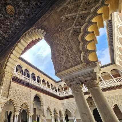 La imagen presenta elementos arquitectónicos encontrados en el Patio de las Doncellas dentro del Alcázar de Sevilla, mostrando componentes esenciales como el arco, el patio, las ventanas de doble arco, el techo de madera, los muros y las columnas, y el cielo que se observa en la fotografía durante el Recorrido Guiado por el Alcázar de Sevilla. La imagen captura detalles característicos de la arquitectura del Alcázar, incluyendo el imponente arco ornamentado con patrones geométricos y arabescos, que conduce a un patio o espacio abierto. Sobre el arco principal, se pueden ver las ventanas de doble arco con balaustradas, típicas de la influencia islámica en la construcción. Otro elemento destacado es el techo de madera con intrincados tallados y un complejo diseño geométrico, que muestra la exquisita artesanía presente en el Alcázar. Los muros y columnas circundantes también están ricamente decorados con motivos similares, creando un juego de luz y sombra que agrega profundidad a la escena. La fotografía está tomada desde un ángulo bajo, lo que permite apreciar la altura y la grandeza de la estructura, con el cielo despejado visible en la parte superior de la imagen. Este enfoque en los detalles arquitectónicos es característico de los recorridos guiados por el Alcázar de Sevilla, que buscan resaltar la riqueza y la importancia de este patrimonio histórico.