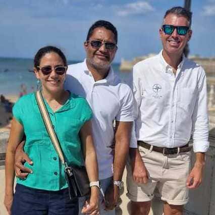 The photo captures three adults standing closely together, smiling, and posing against the backdrop of the beach. On the left is a woman in a vibrant green top, sporting sunglasses and carrying a shoulder bag. In the center stands a man in a white polo shirt with sunglasses casually draped on the collar, while on the right is the guide from Check-In Sevilla, dressed in a white shirt with a nautical-themed logo. All three individuals don shorts, exuding a relaxed and cheerful demeanor. The clear blue sky indicates a sunny day, and the beach, with a few visitors, suggests a tranquil setting ideal for a Seville Private Tours The distant view of the ocean enhances the picturesque scene