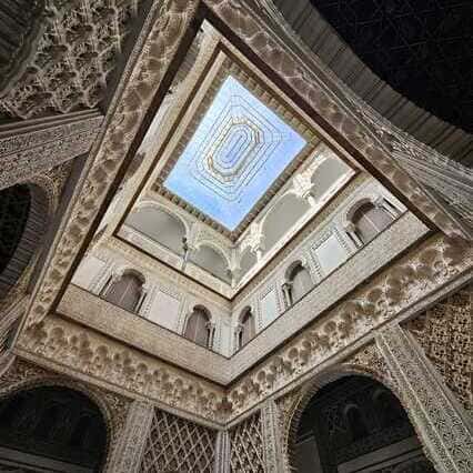 Una vista hacia arriba del Patio de las Muñecas en el Alcázar de Sevilla, resaltando sus detallados trabajos de yeso mudéjares, patrones geométricos y ventanas arqueadas, todo enmarcado por un cielo azul despejado. Explora esta maravilla arquitectónica durante el Recorrido Guiado por el Alcázar de Sevilla. La imagen captura la belleza y la riqueza de detalles del Patio de las Muñecas, un espacio que refleja la influencia islámica en la arquitectura del Alcázar de Sevilla. Los intrincados trabajos de yeso, los diseños geométricos y las ventanas arqueadas son elementos clave que definen el estilo mudéjar de esta sección del palacio. Esta vista panorámica desde el interior del patio invita a los visitantes a apreciar la maestría artesanal y la armonía que se logra entre la arquitectura y el cielo despejado que la enmarca. Durante el recorrido guiado por el Alcázar, los visitantes tendrán la oportunidad de explorar y descubrir de cerca esta maravilla arquitectónica, sumergirse en su historia y comprender la riqueza cultural que la define. La imagen evoca la sensación de asombro y admiración que puede experimentar el visitante al contemplar la belleza y la complejidad del Patio de las Muñecas, un espacio que representa la fusión de estilos y la herencia artística del Alcázar de Sevilla.