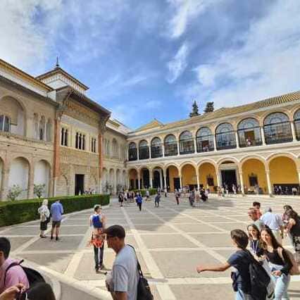 Un bullicioso patio con visitantes en el Alcázar de Sevilla, que presenta un edificio de dos pisos con arcos amarillos en el nivel inferior y una serie de ventanas en el nivel superior, todo bajo un cielo azul. Explora este patio durante el Recorrido Guiado por el Alcázar de Sevilla. La imagen captura la actividad y la energía del patio del Alcázar de Sevilla, donde los visitantes se mueven entre los elementos arquitectónicos característicos de este espacio. Los arcos amarillos en la planta baja y las ventanas en el segundo nivel crean una composición visual atractiva, enmarcada por un cielo despejado. Durante el recorrido guiado por el Alcázar, los visitantes tendrán la oportunidad de explorar este patio y sumergirse en la atmósfera vibrante y llena de vida que lo caracteriza. Podrán apreciar de cerca la belleza de la arquitectura, con sus detalles y elementos distintivos, y comprender mejor la importancia de este sitio histórico. La imagen invita a los espectadores a imaginar la experiencia de visitar el Alcázar de Sevilla y descubrir los diversos patios y espacios que conforman este impresionante conjunto arquitectónico. La guía durante el recorrido permitirá a los visitantes apreciar en profundidad la riqueza y la historia de este lugar emblemático.