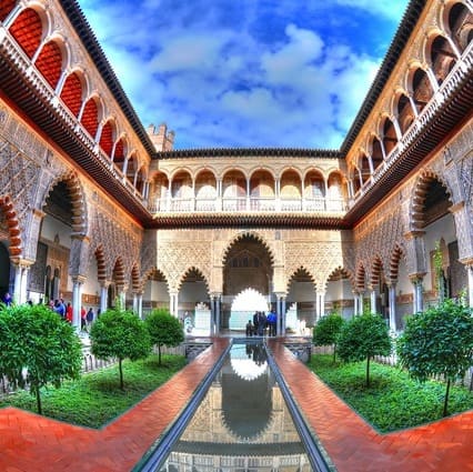 Una imagen del Patio de las Doncellas con un estanque central de reflexión, rodeado de ornamentados arcos, columnas y plantas verdes bajo un brillante cielo azul. Explora esta joya arquitectónica durante el Recorrido Guiado por el Alcázar de Sevilla. La imagen captura la belleza y la elegancia del Patio de las Doncellas, un espacio emblemático del Alcázar de Sevilla. El estanque central, rodeado por una composición simétrica de arcos decorativos y columnas, crea un entorno sereno y armónico, acentuado por la presencia de la exuberante vegetación y el cielo despejado. Durante el recorrido guiado por el Alcázar, los visitantes tendrán la oportunidad de explorar de cerca este patio y sumergirse en su riqueza arquitectónica y cultural. Podrán apreciar los detalles y la maestría de los elementos constructivos, comprendiendo la importancia de este espacio como una joya del patrimonio sevillano. La imagen invita a los espectadores a imaginar la experiencia de visitar el Patio de las Doncellas, donde la arquitectura, el agua y la naturaleza se funden en una composición visual cautivadora. El recorrido guiado les permitirá descubrir los significados y las historias que se esconden detrás de este impresionante conjunto arquitectónico.