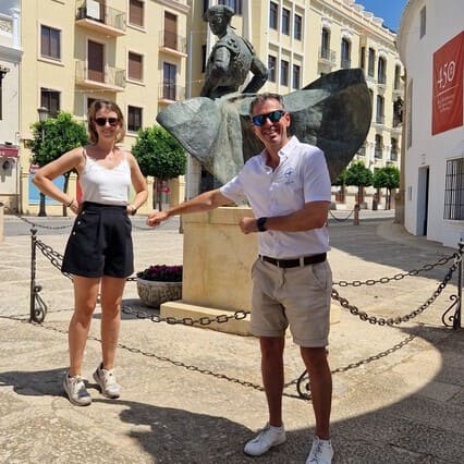 The image captures two individuals, the guide from Check-In Sevilla and a woman, positioned on either side of a bronze sculpture of a bullfighter at the main gate of the Ronda Bullring. The dynamic sculpture portrays the bullfighter in action, with a dramatic cape unfurled behind him. The man and woman playfully mimic the pose, creating a memorable photo opportunity that showcases Ronda's rich bullfighting heritage. The clear skies and sunny day enhance the picturesque setting of the plaza, featuring classical architectural elements and ornamental plants. This scene epitomizes the historical allure and cultural significance of bullfighting in Ronda during one of our Seville Private Tours.